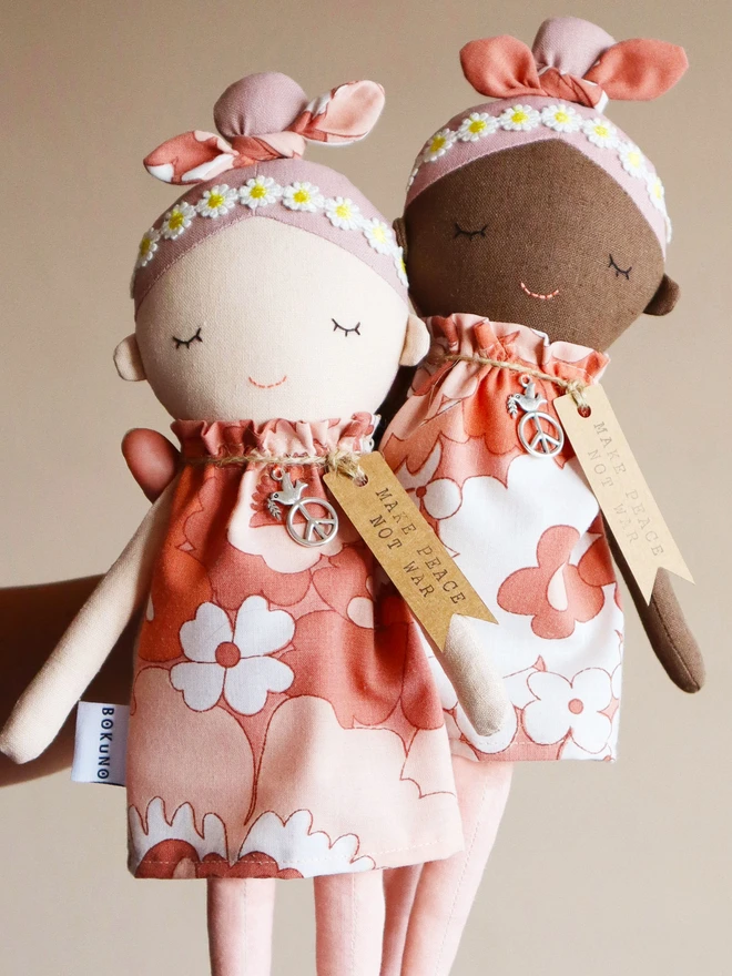 Two handmade fabric dolls called Peace Wishing Flower Child Dolls, each wearing peach and white retro floral dresses, daisy headbands, and bow topknots. They have serene embroidered faces, peace sign charms around their necks, and tags reading “Make Peace Not War.” One doll has a light complexion and the other has a darker complexion, both held side by side against a plain background.