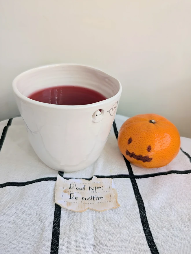 Small ghost cups with finger dents in white handmade ceramic with a tiny sculpted ghost in a cut out near the top of the beaker with a hand painted bubble with the word boo