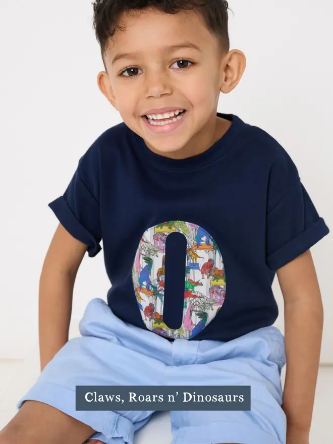 a 4 year old boy wearing a personalised navy t-shirt with his initial cut from liberty print featuring dinosaurs