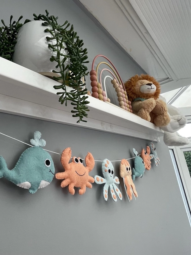 Nursery Under The Sea Garland, a sea themed garland hanging from a white shelf against a grey backdrop in a child's bedroom. 