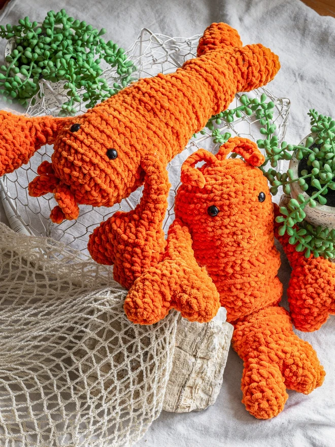 Chunky Chenille Cuddly Crochet Crustacean Lobster Plush, an orange lobster plush sitting next to another lobster on a white surface accompanied by a bag and some greenery. 