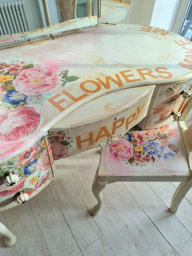 An antique kidney-shaped dressing table with chair and a trio of mirrors. Upcycled in cream and pale pink with decoupage florals in oranges and pinks. Text across the table read ‘flowers make me happy’ in the orange of the undercoat.