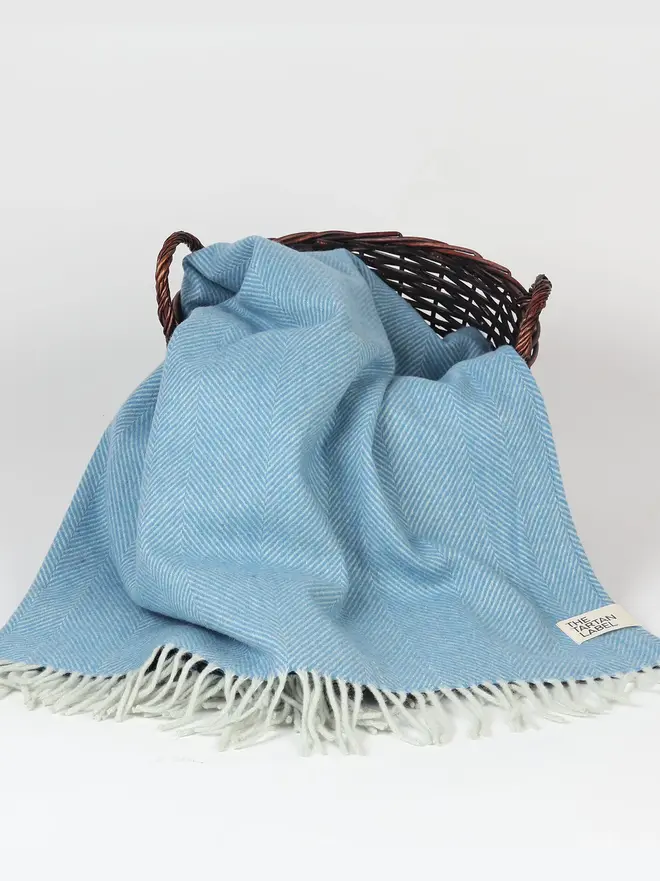 Sky Recycled Wool Tartan Blanket, a blue picnic blanket sitting on a wooden basket on a white surface in front of a plain backdrop. 