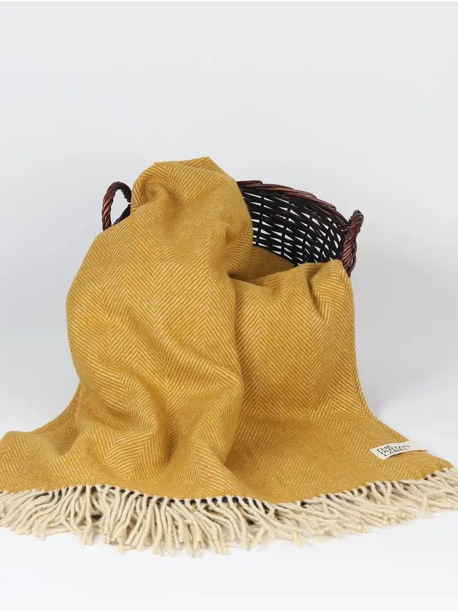 Mustard Recycled Wool Tartan Blanket, a yellow picnic blanket sitting on a wooden basket on a white surface in front of a plain backdrop. 
