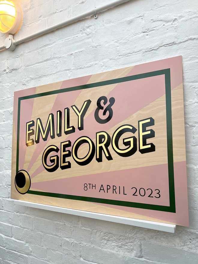 Modern wedding sign featuring the couple's names in gold leaf, outlined in black, and the wedding date, with a sunburst background and a border. Shown against a white brick wall, at an angle. 