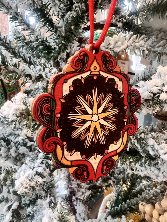 Multi coloured wooden tree decoration with a star pattern at the centre. It is hanging by a red ribbon on a fake white christmas tree with other decorations. 