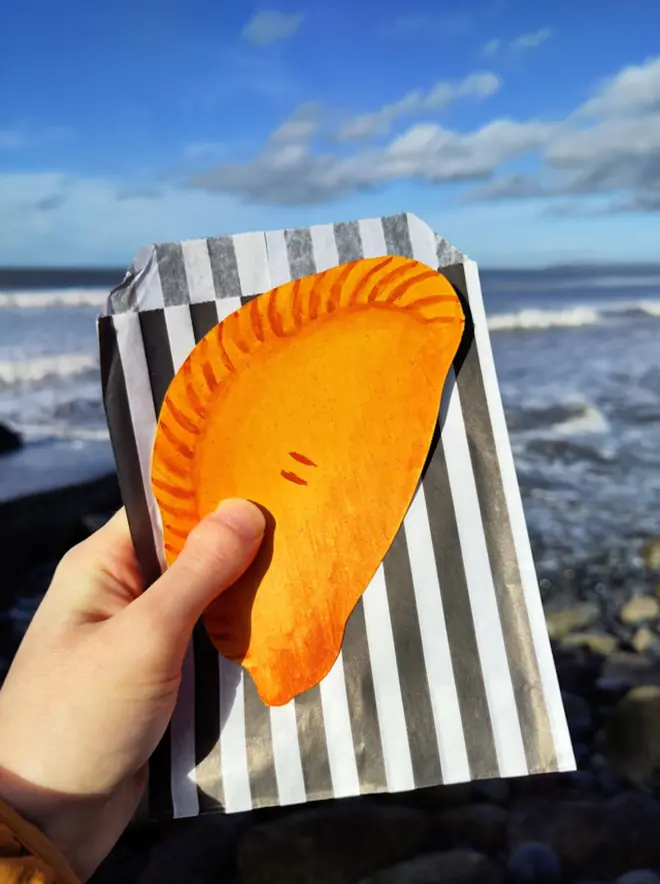 hand painted cornish pasty drawing, with black and white striped paper bag, held in front of ocean view