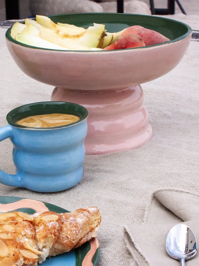 a colourful pedestal bowl filled with fruit. the bowl has a wide shallow bowl and a chubby elevated foot. The bowl is blush pink on the outside and dark forest green on the inside. The bowl is sitting on a patio table surrounded by a tablecloth and breakfast food and matching coffee cups