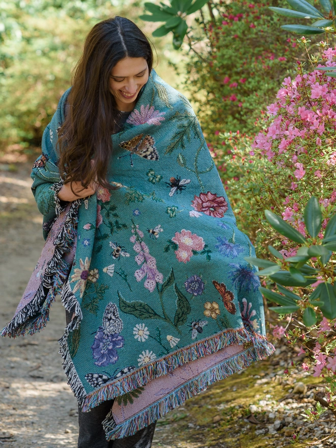 A woman seen wrapped in a 'Pollination Bloom' Recycled Cotton Blanket walking outside.