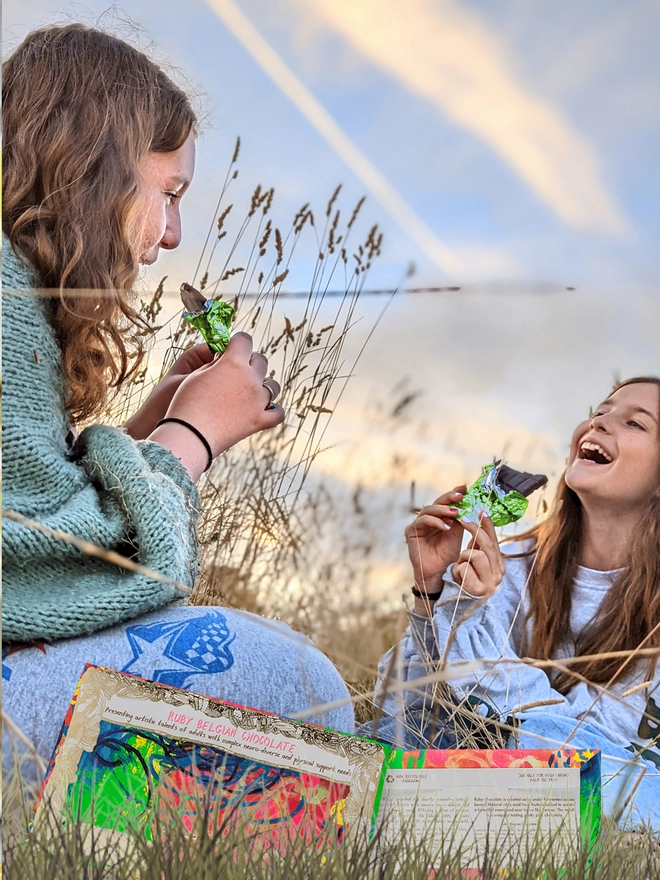 Two happy girls in a field enjoying charity ruby chocolate bar wrapped in green foil 