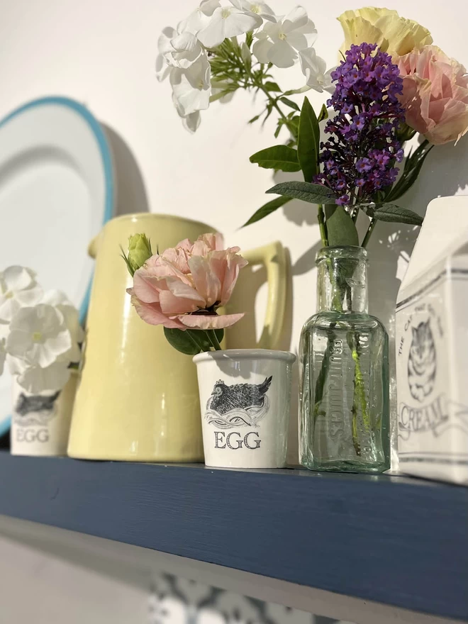 On a shelf with jugs and plates, stand two handmade ceramic egg cups each holding a flower.
