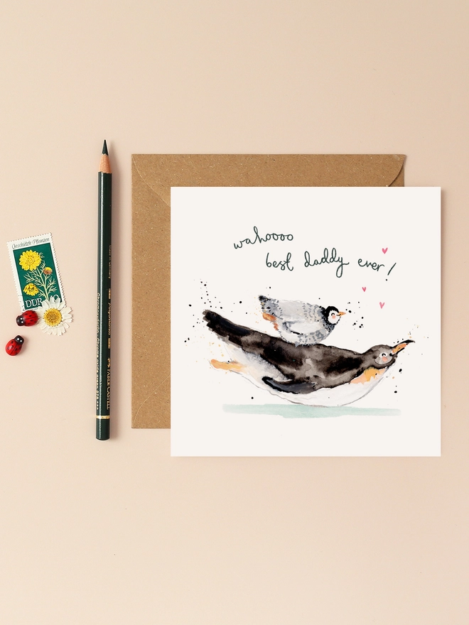 A charming illustration of a father penguin carrying a young penguin on its back, both joyfully surrounded by scattered hearts. The playful handwritten message "Wahoo Best Daddy Ever!" adds a heartwarming touch.