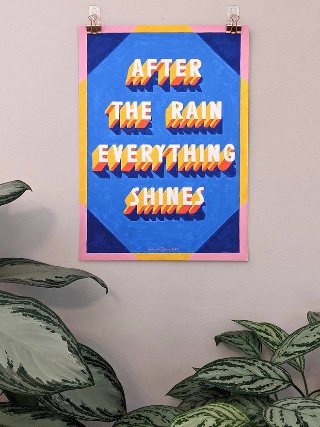 Art print of painted 3d typography reading After The Rain Everything Shines hanging with clips on a wall with plants below