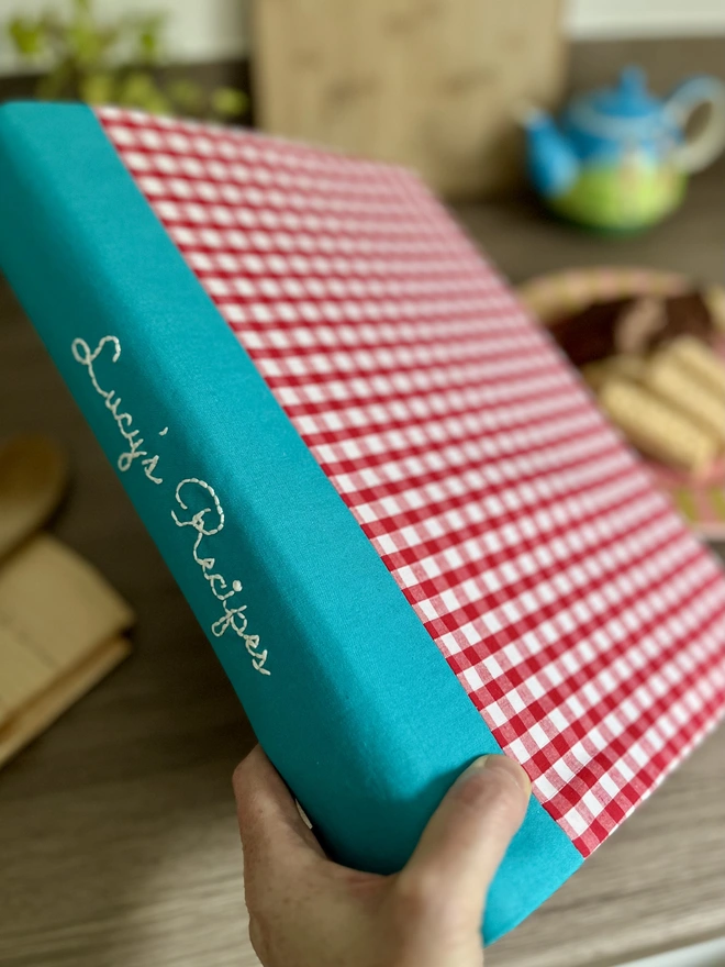 Model's hand holds an A4 ring binder with a red and white gingham fabric cover, angled to show personalisation on the teal spine, with a kitchen bench in the background where afternoon tea waits.
