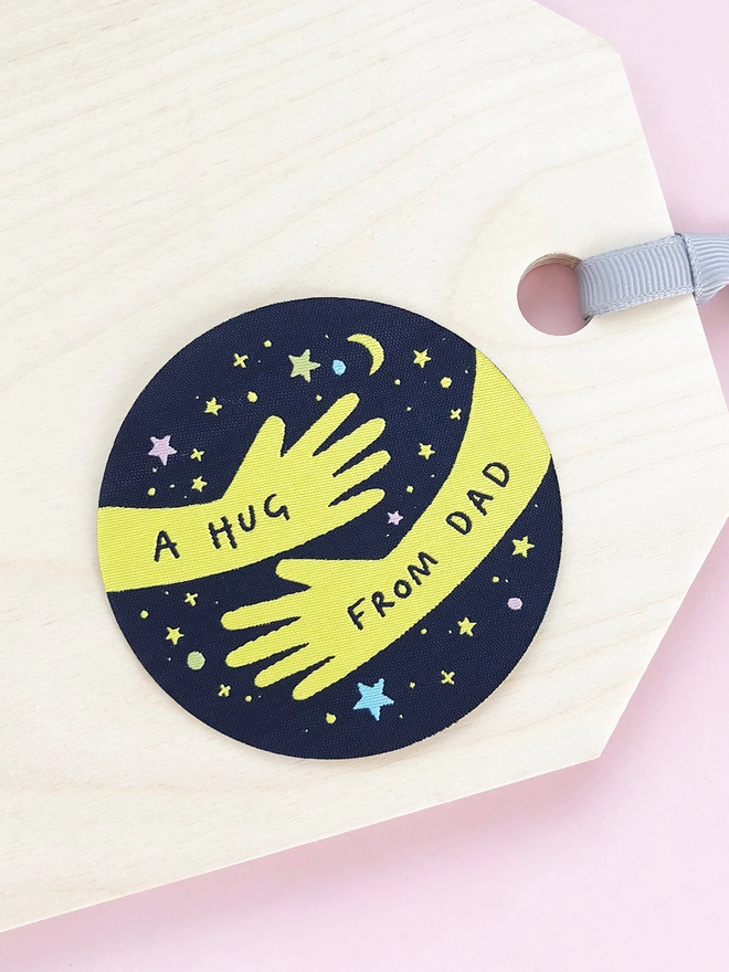 A navy blue and yellow woven patch with two arms in a hugging position and the words "A hug from Dad" lays on a wooden desk.