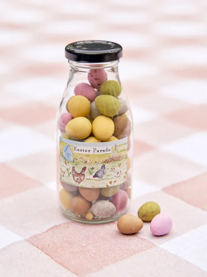 Glass bottle with a black lid, filled with multi coloured chocolate eggs on a pink checkered tablecloth
