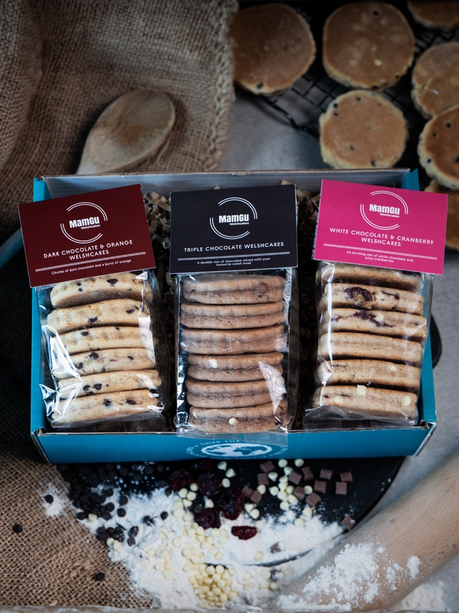 3 packs of chocolate flavoured Welsh Cakes in a gift box on a busy kitchen work top with welsh cakes on a cooling rack, flour, chocolaten chips, rolling pin and wooden spoon