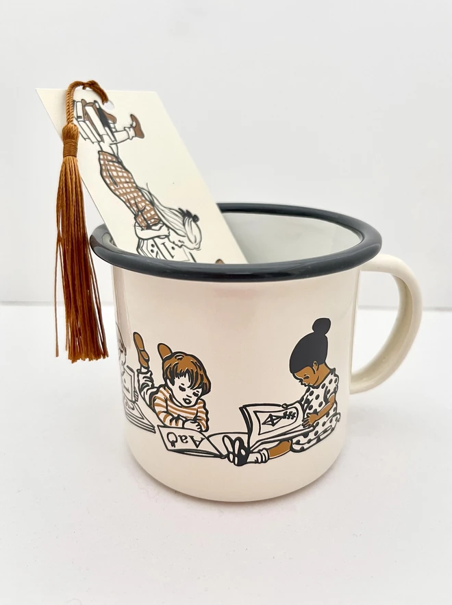 an enamel mug cream in colour with a dark grey rim featuring a 2 colour screen print of children reading books. Inside the mug is a matching bookmark