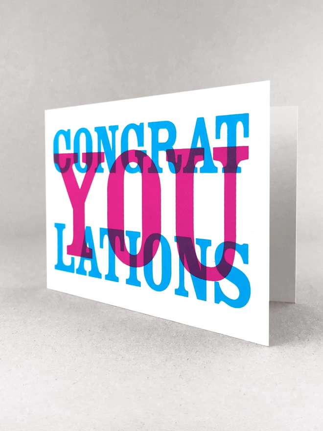 This landscape card reads Congrat YOU lations printed in cyan and magenta in overlapping colours. Stood slightly open, in a light grey studio set.