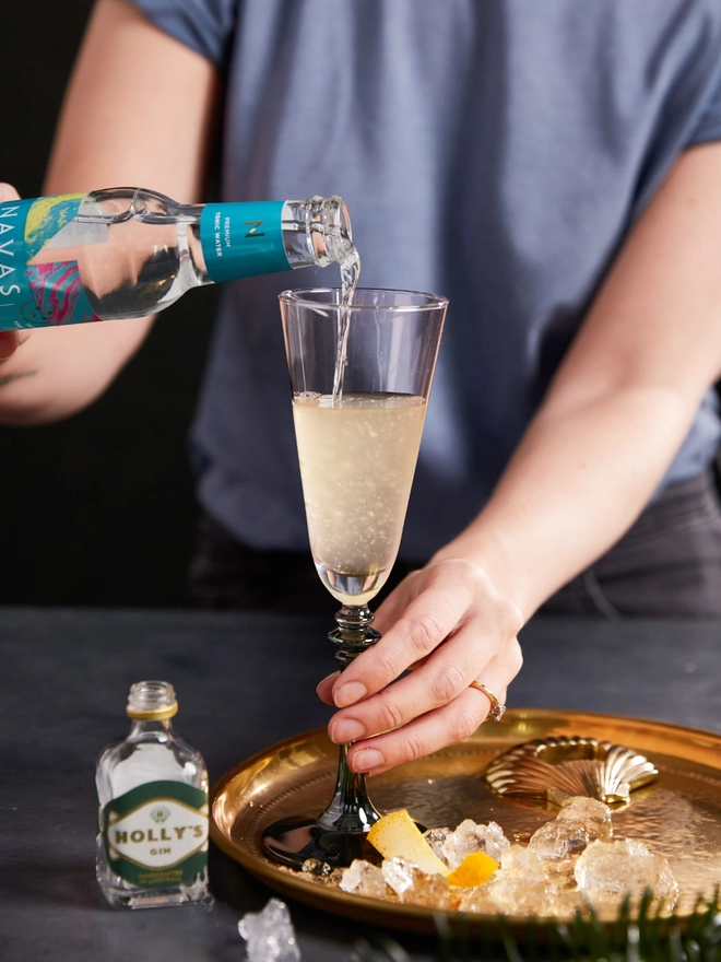 Pouring Tonic water into a cocktail glass with Holly's Gin miniature on the table