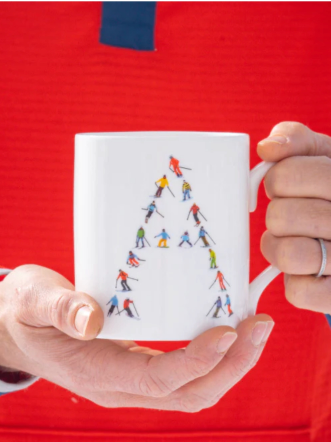 A simple, white mug illustrated with an ‘A’ made of small, multicoloured skiers. The mug is held in two hands and is set against the background of a contrasting, red torso.