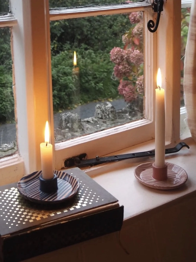 Ceramic Patterned Candlestick Holder, a pair of colourful candlestick holders sitting on a window sill. They are accompanied by a lit candle. 
