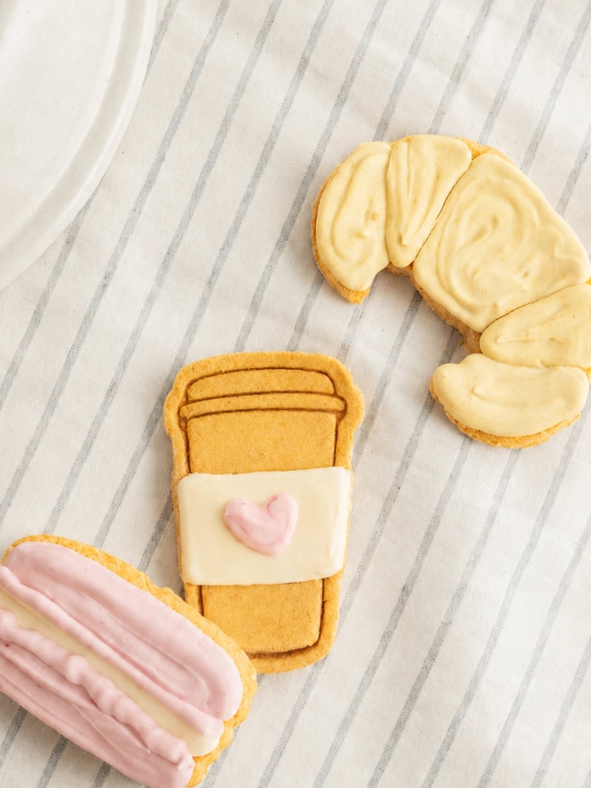 Parisian Bakery iced dog biscuit trio styled on striped fabric beside a ceramic plate.