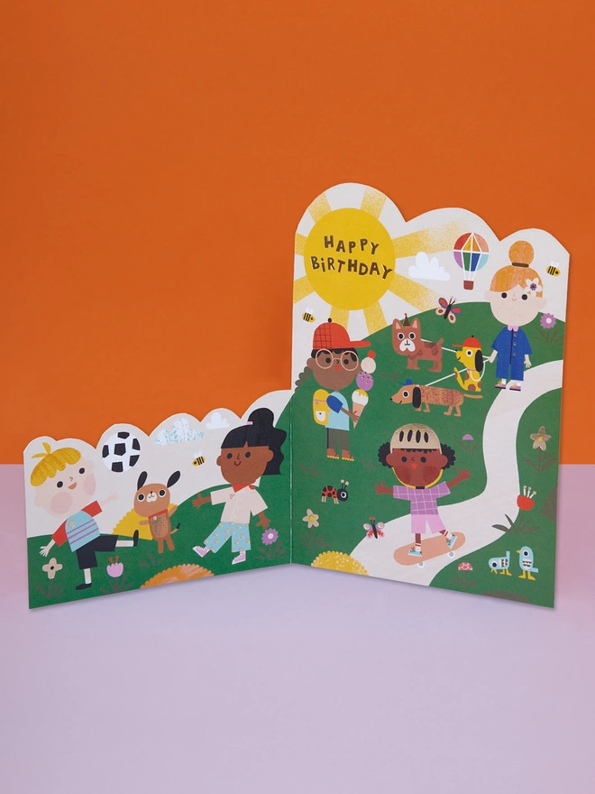 A sunny day at the park themed, die cut children’s birthday card, filled with colour and fun characters skateboarding, playing football, walking their dogs, eating ice cream and enjoying their day at the park. The card is complete with gold foil details and a gold foil ‘Happy Birthday’ message