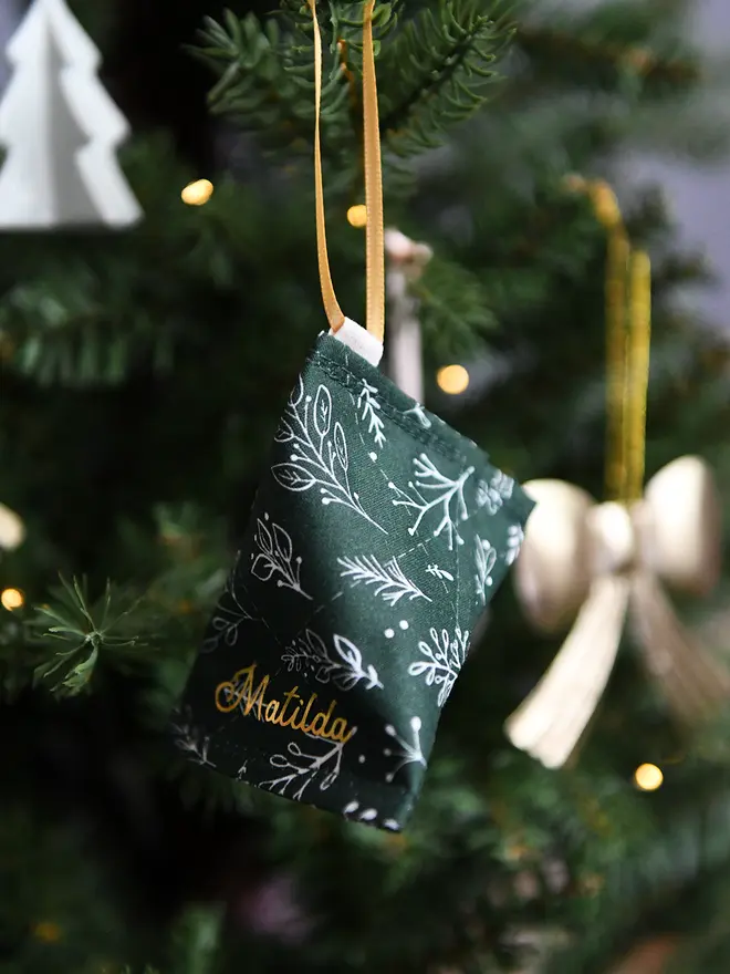 A handmade fabric fortune cookie Christmas decoration in green festive foliage print, personalised with the name “Matilda” in gold, hangs from a golden ribbon on a Christmas tree branch.