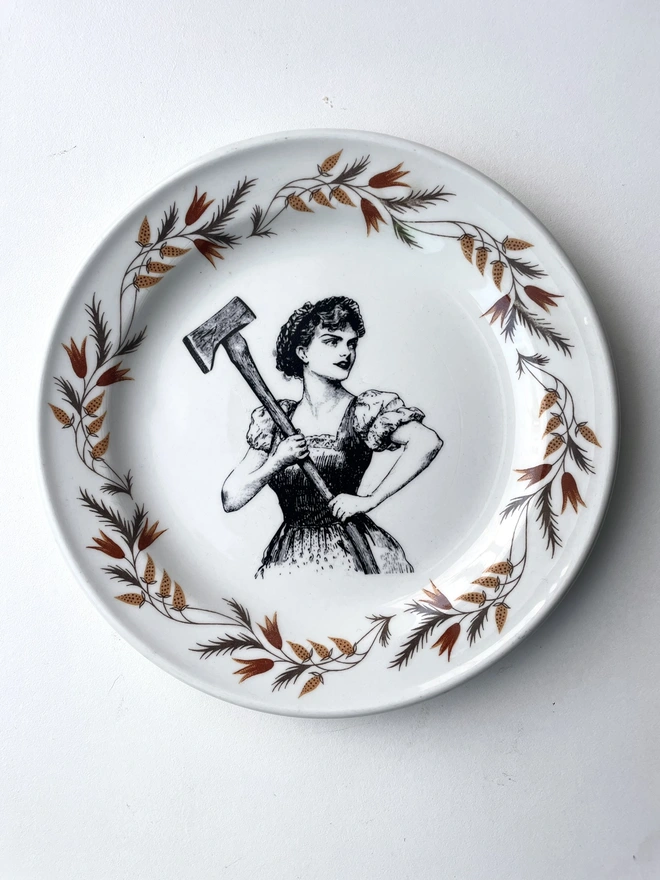 A vintage plate with a patterned border , in the centre of the plate is a black and white vintage illustration of a beautiful lady in a floral dress wielding an axe 