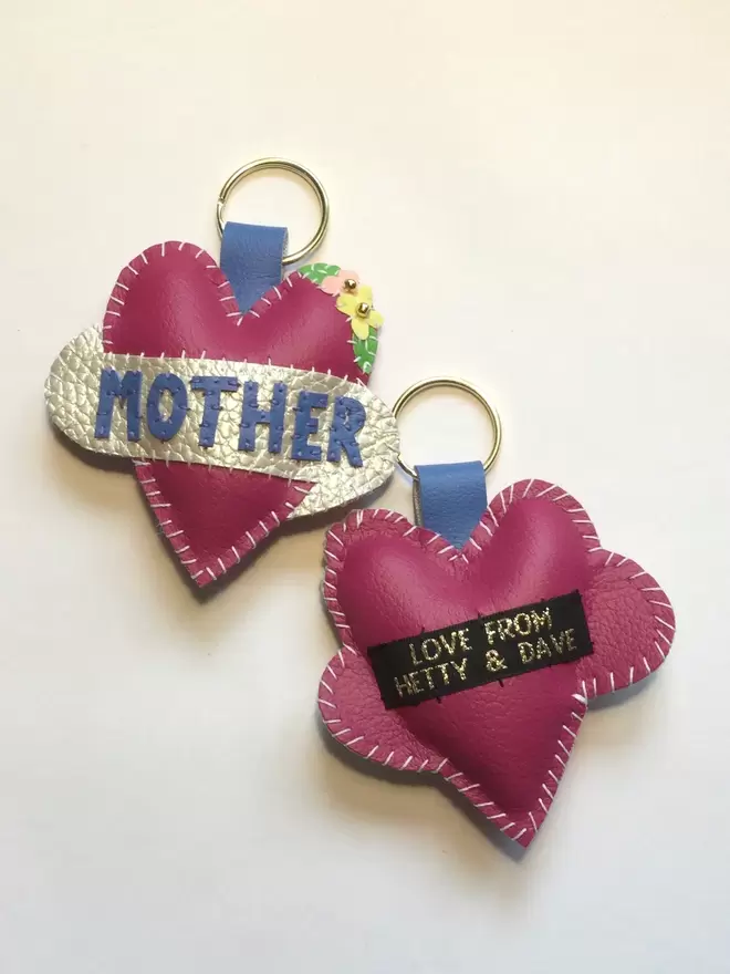 Two pink heart keyrings. One spells MOTHER in blue lettering on a white scroll, the other shows the plain pink reverse with a black Love From Hetty & Dave label