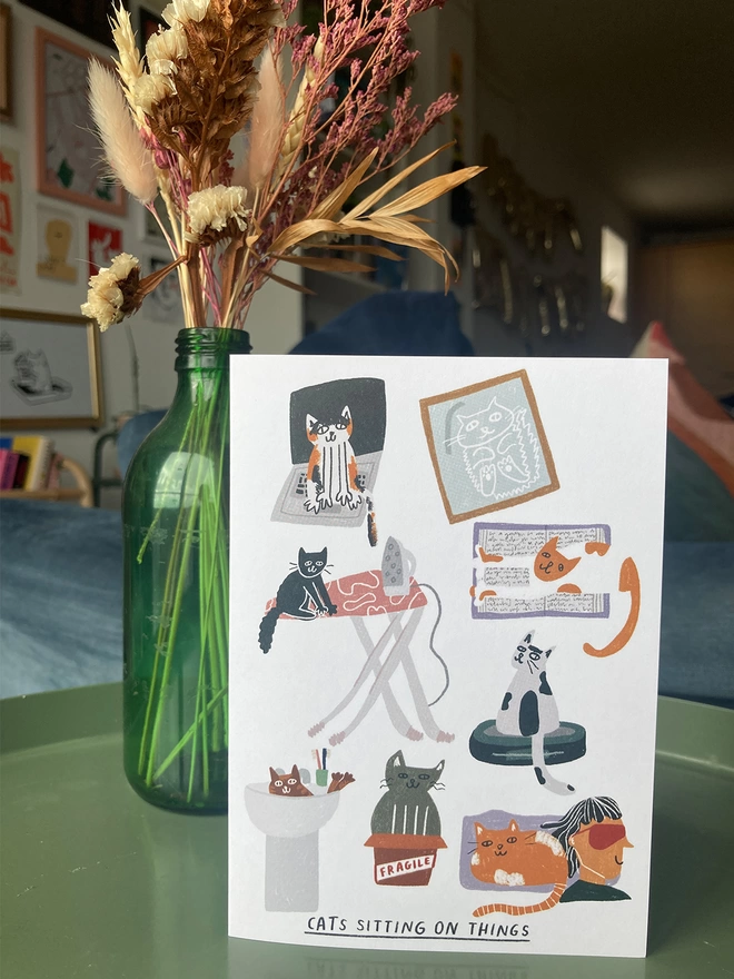 Greetings card with illustrations of cats sitting on things. Placed on a table with flowers in the background.