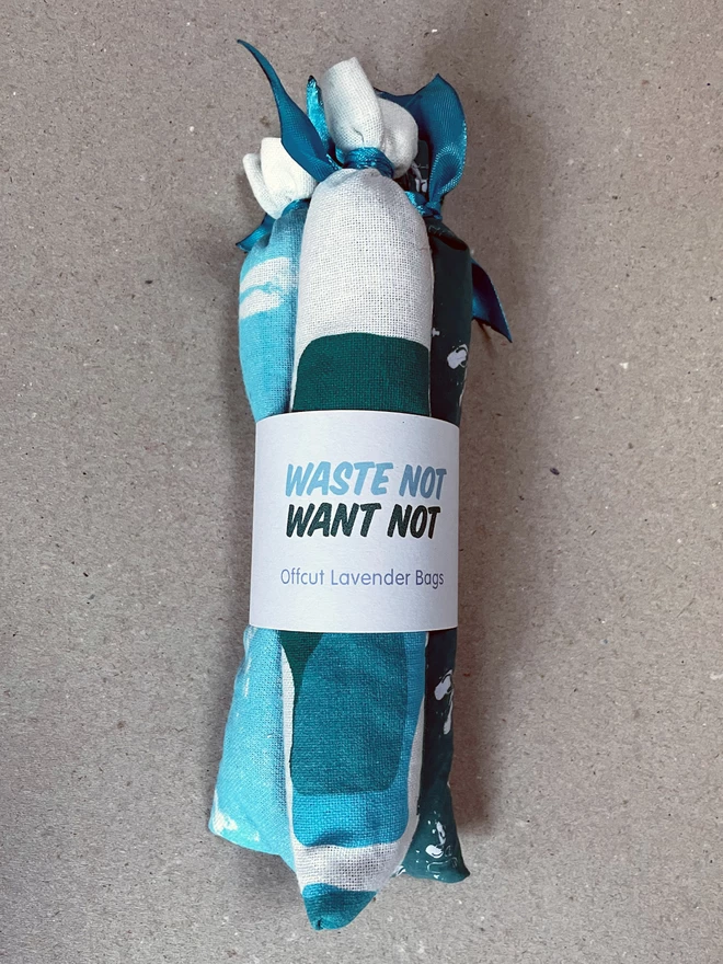 A bundle of three long style cotton lavender bags, tied at the tops, gathered with a branded cuff, sit on a greyboard background - it says Waste Not Want Not, Offcut Lavender Bags. The fabric has bits of turquoise and teal pattern visible.