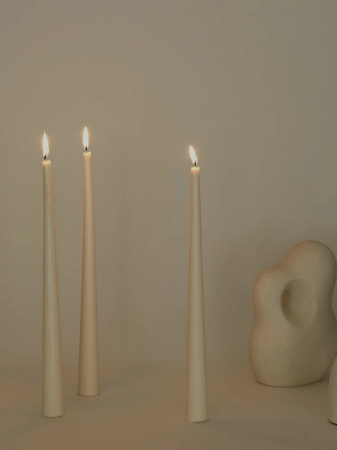 White Stem Candles (Set Of 3). Three lit cream taper candles with tapered bases, standing beside sculptural speckled stoneware ornaments against a soft neutral wall. The elegant, self-standing candles bring a calm, warm glow to modern living spaces.