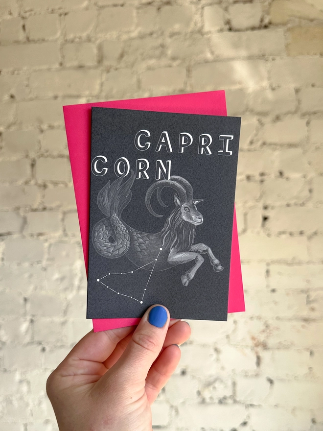 a greetings card featuring a greyscale illustration of a zodiac style sea goat with the text “CAPRICORN” with a neon pink envelope