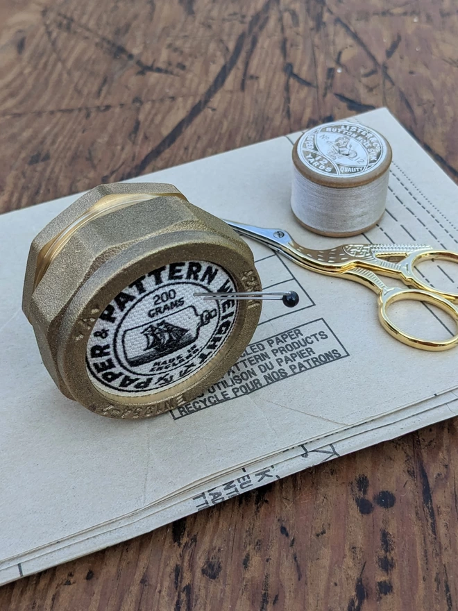 Black pattern and paper weight on its side holding a needle and pin on vintage sewing pattern paper next to embroidery scissors and cotton reel