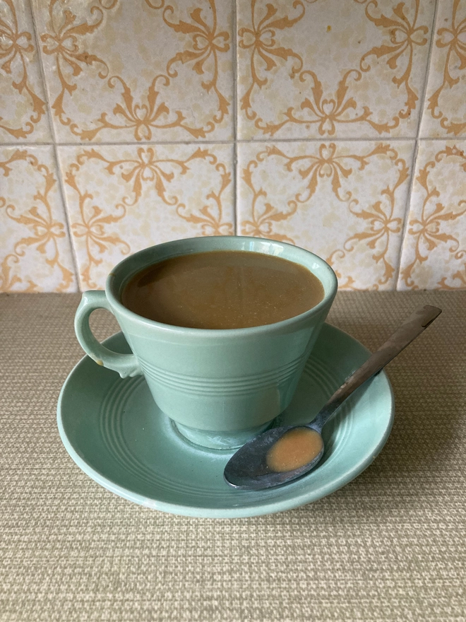 A pale green 1940s cup and saucer from the company Beryl Ware. The cup is filled to the brim with resin 'tea'. There is a tea spoon in the saucer which has a drop of resin tea in its bowl.