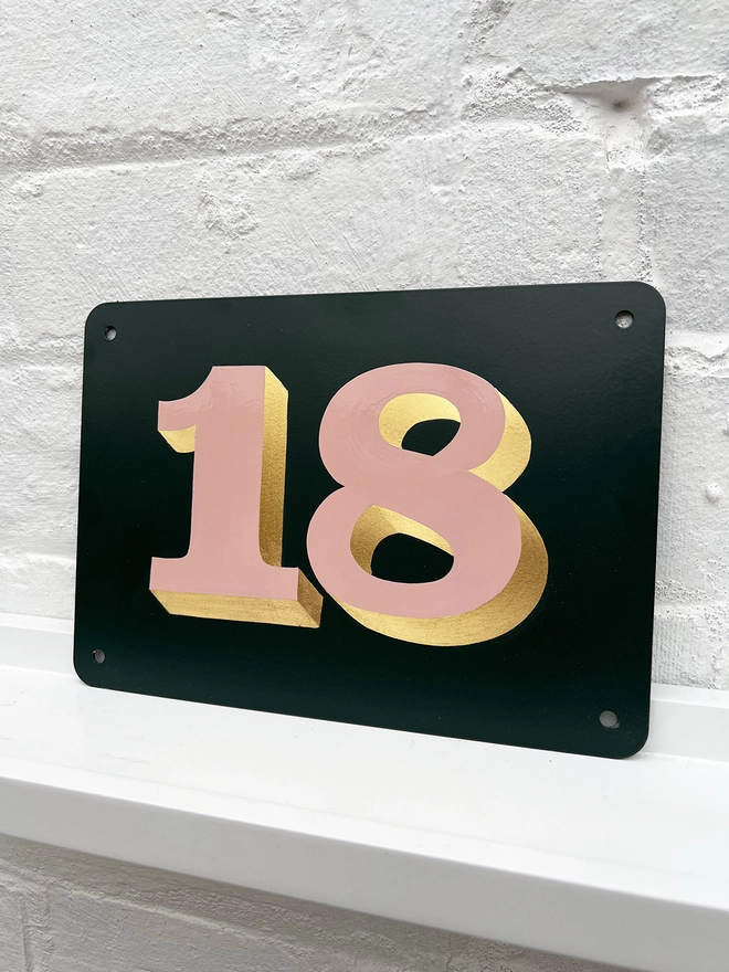 Hand painted house number against a white brick wall, number 18 in serif style in dusky pink and directionally burnished 23 carat gold leaf, on anthracite grey background. 
