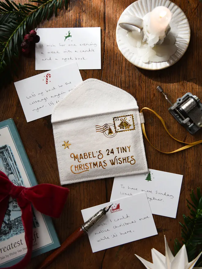 A handmade felt envelope advent calendar with golden lettering that reads 'Mabel's 24 Tiny Christmas Wishes' with several Christmas notecards all laying on a wooden table.