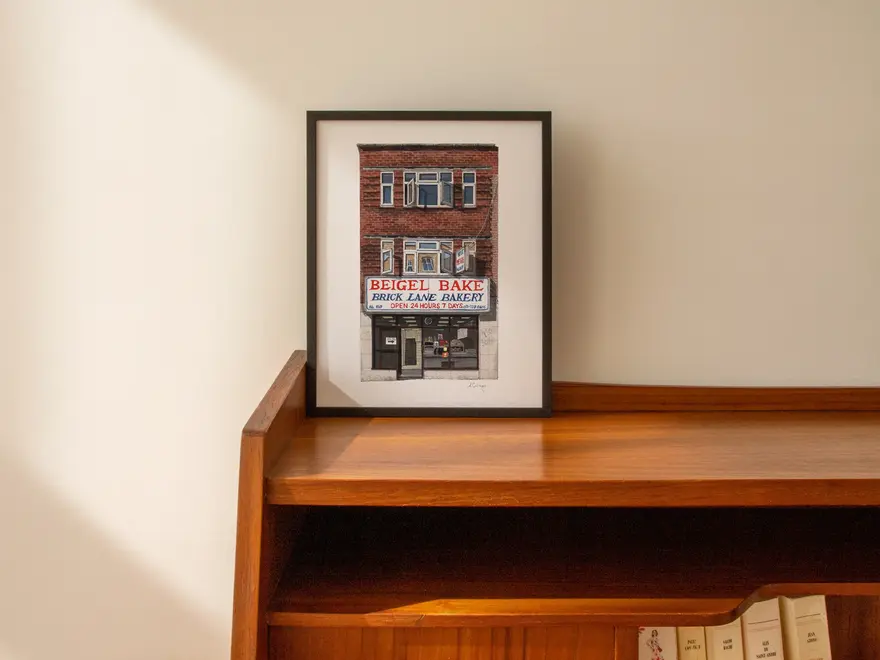 Brick Lane Beigel Bake Giclée Print (Unframed) Shoreditch, a print of a cafe sitting in a black frame on a wooden surface against a plain backdrop. 