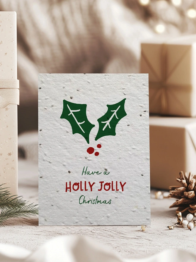 Christmas card with a holly leaf on with 'Have a Holly Jolly Christmas'. Behind the card is some presents, a pine cone and pine leaves.