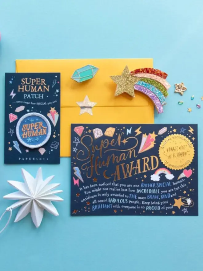 The navy award certificate lies with the patch card on a yellow envelope. A rainbow glitter shooting star, an enamel crystal pin and a white paper star complete the scene.
