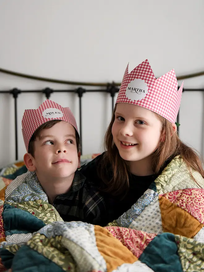 Two children wearing red gingham personalised Christmas crowns are wrapped in a cosy patchwork quilt giggling together.