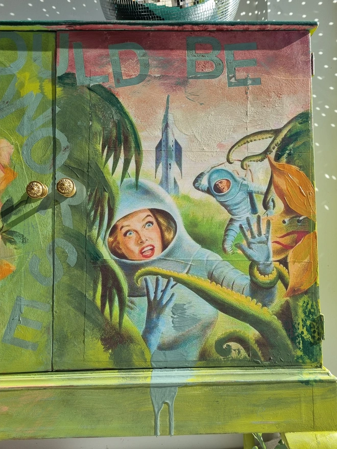 A painted record cabinet decorated with a sci fi image from the 1950s of a lady running from an alien and on the other door, a rose taken from a Victorian seed packet. There is wording that runs down the middle of the cabinet which reads 'it could be worse'. The colours are apple green and pinks.