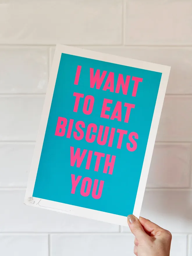 'I Want to Eat Biscuits with You' screen print in bright blue with neon pink text, held up against a white tiled wall