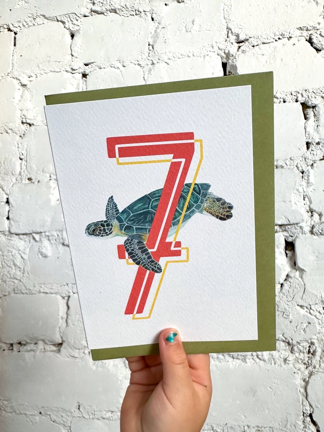 a white card with a big red number 7 and an illustrated green sea turtle with a green envelope