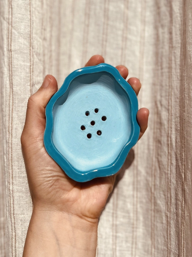 wavy rimmed soap dish held in the palm of a hand against a fabric background. the soap dish is bright blue on the outside with a sky blue on the inside