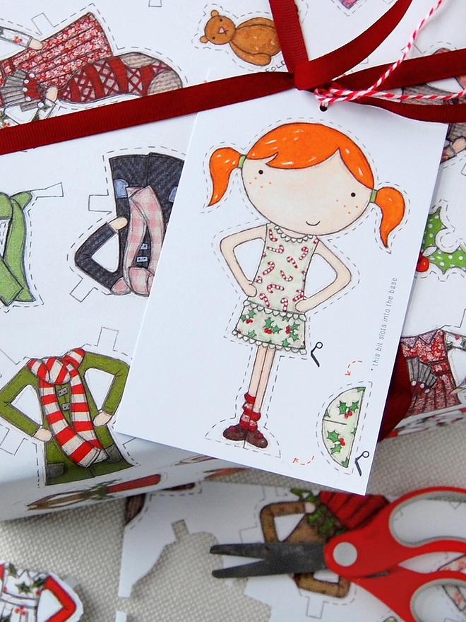 A gift wrapped in Christmas wrapping paper with illustrated outfits for the included paper doll tag.