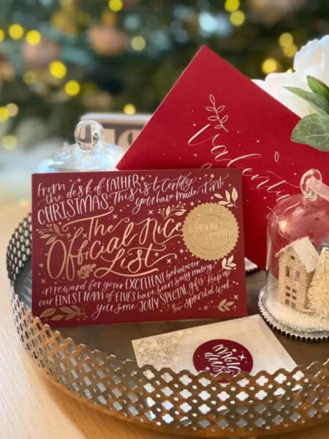 A red gold foil certificate and hand-lettered envelope on a silver tray by twinkling tree lights.