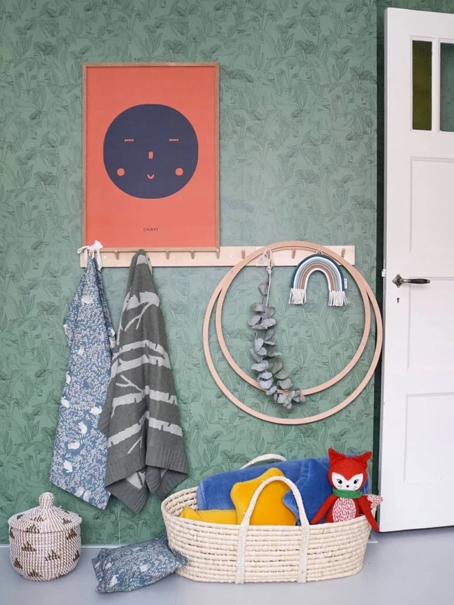 A picture of some hanging wall hooks mounted on a green wall. The birch forest baby blanket hangs from one of the pegd, alongside some wooden hoops and above a moses basket with toys in.